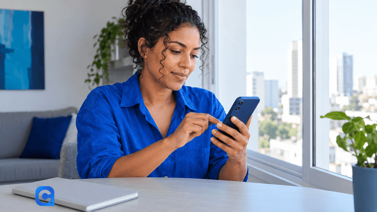 Mulher de blusa azul consulta o celular em uma mesa de casa.