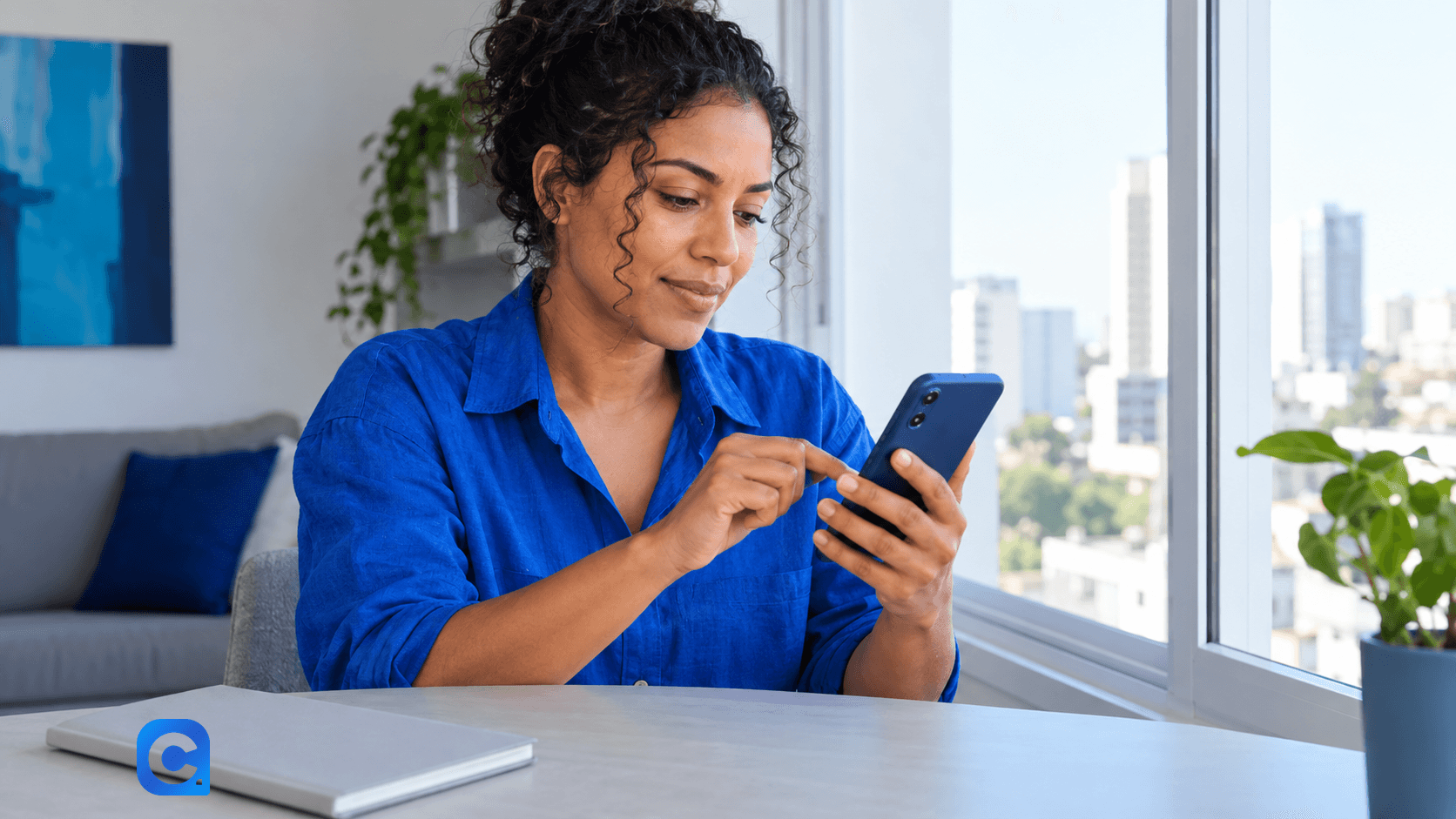 Mulher de blusa azul consulta o celular em uma mesa de casa.